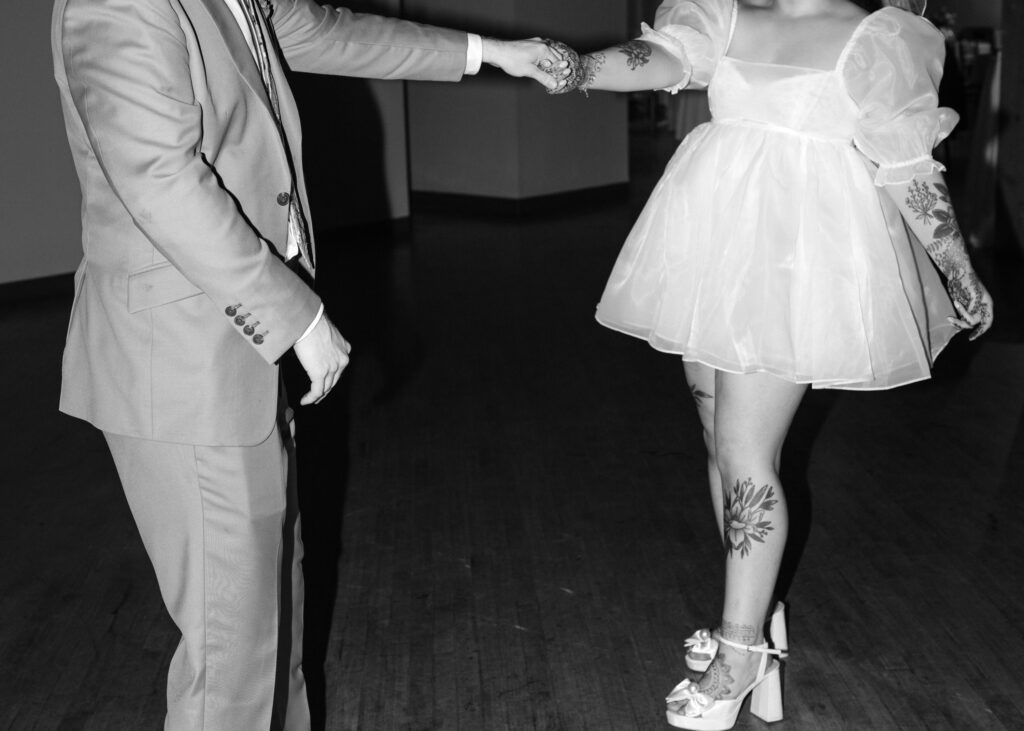 Black-and-white photo of the bride and groom holding hands while dancing during their wedding reception.