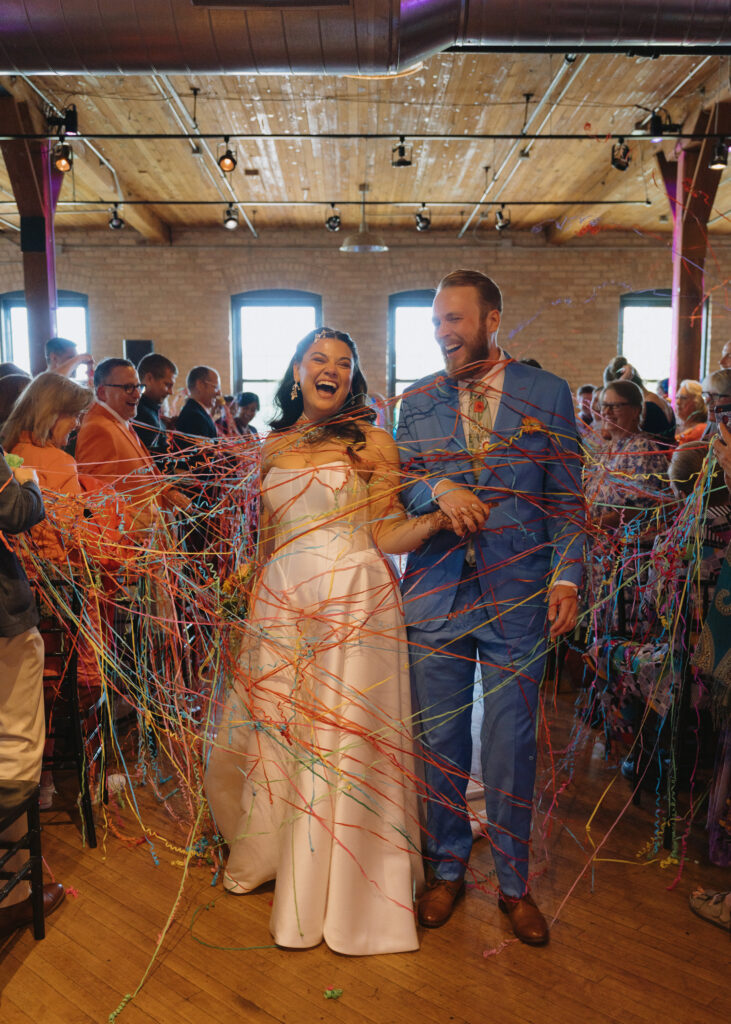 Newlyweds laugh as they walk back down the aisle through colorful ribbon streamers after their wedding ceremony.