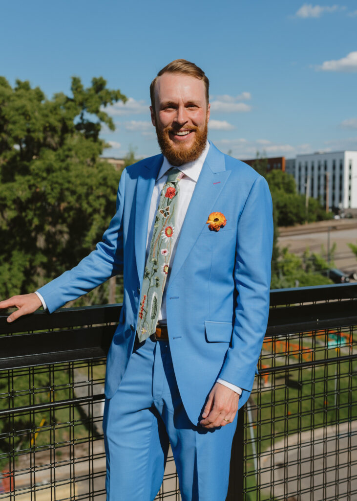 Groom smiles while standing on a rooftop terrace in his bright blue wedding suit.