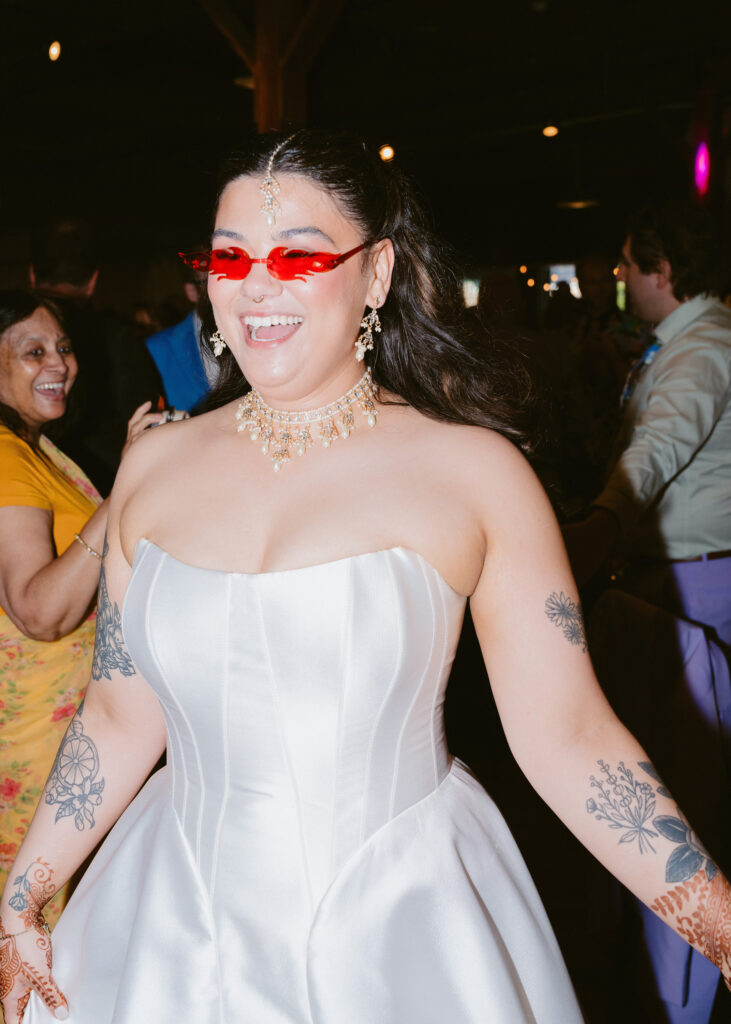 Bride laughs while wearing red party sunglasses on the dance floor during the wedding reception