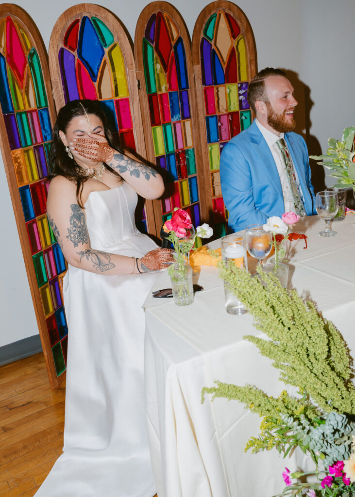 Bride laughs while sitting beside the groom at their sweetheart table during the wedding reception.