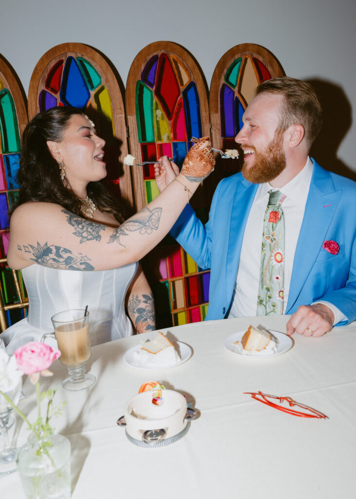 Bride and groom laugh as they feed each other wedding cake during their colorful reception celebration.