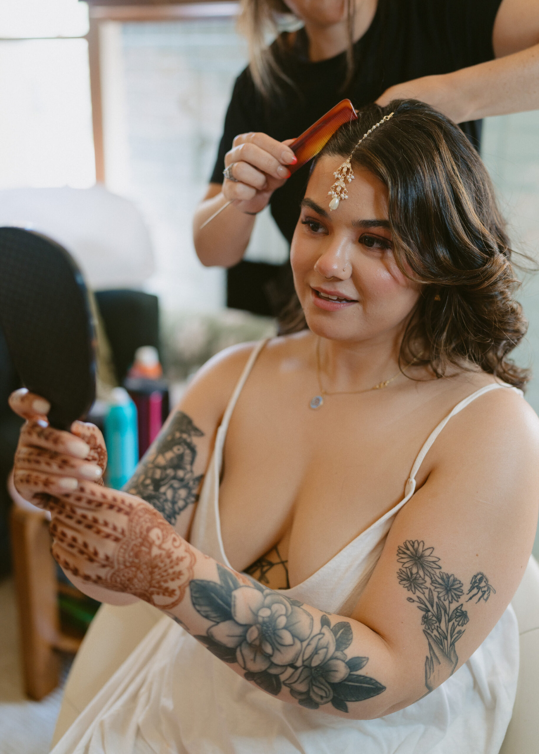 Bride smiles while looking into a handheld mirror as her hairstylist finishes styling her hair during wedding morning preparations.